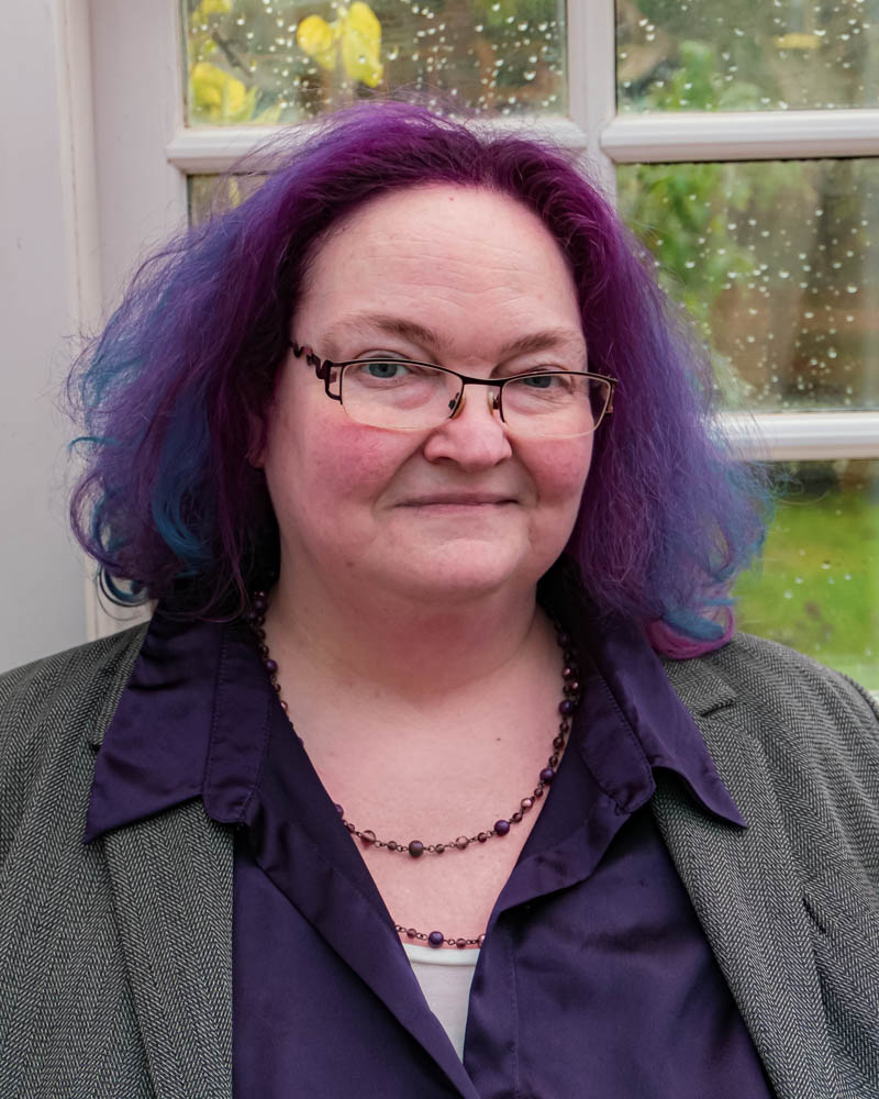 Head and shoulders photo of Sam Griffin smiling. She is a plump middle aged white woman, smartly dressed and wearing glasses. Her hair is vibrant purple.
