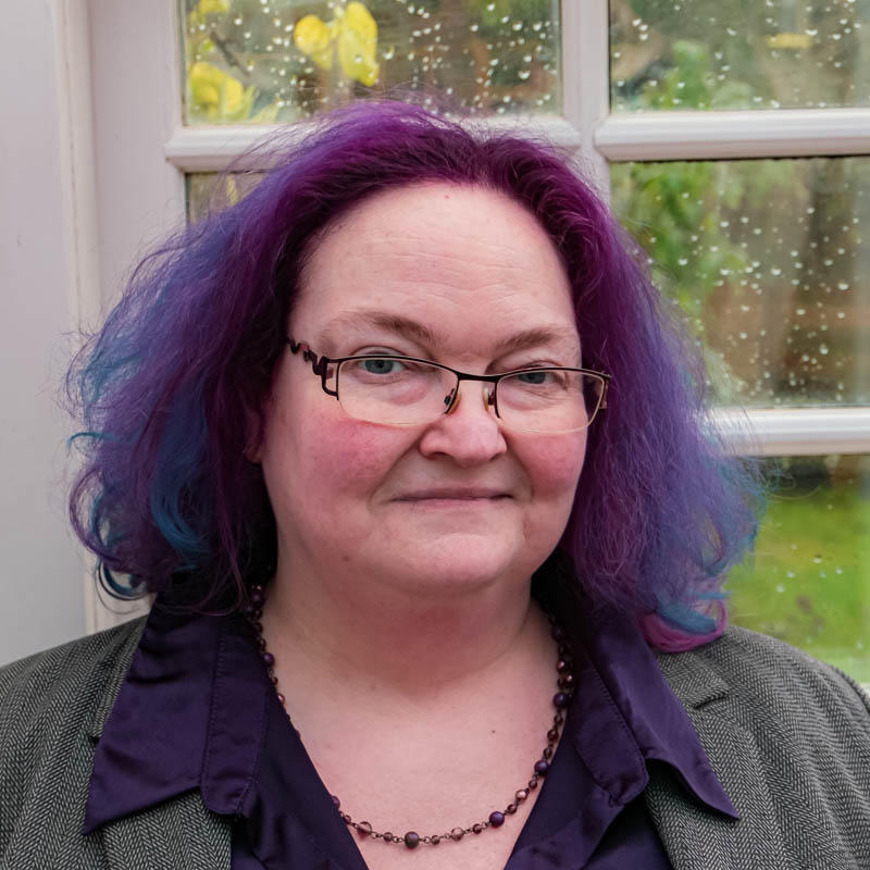 Head and shoulders photo of Sam Griffin smiling. She is a plump middle aged white woman, smartly dressed and wearing glasses. Her hair is vibrant purple.